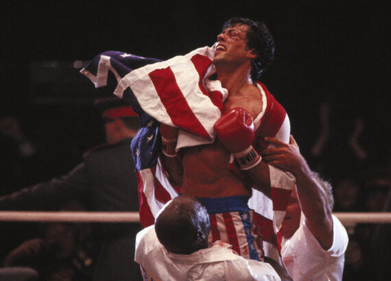 Rocky Balboa wearing an American flag and being held up by two men after a fight