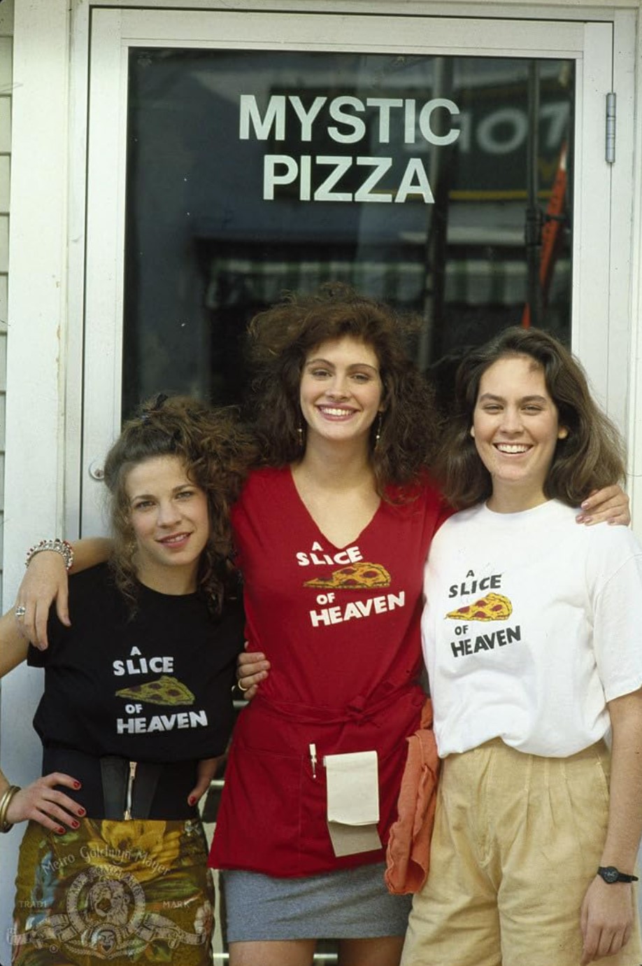 Three women standing in front of a door that says Mystic Pizza