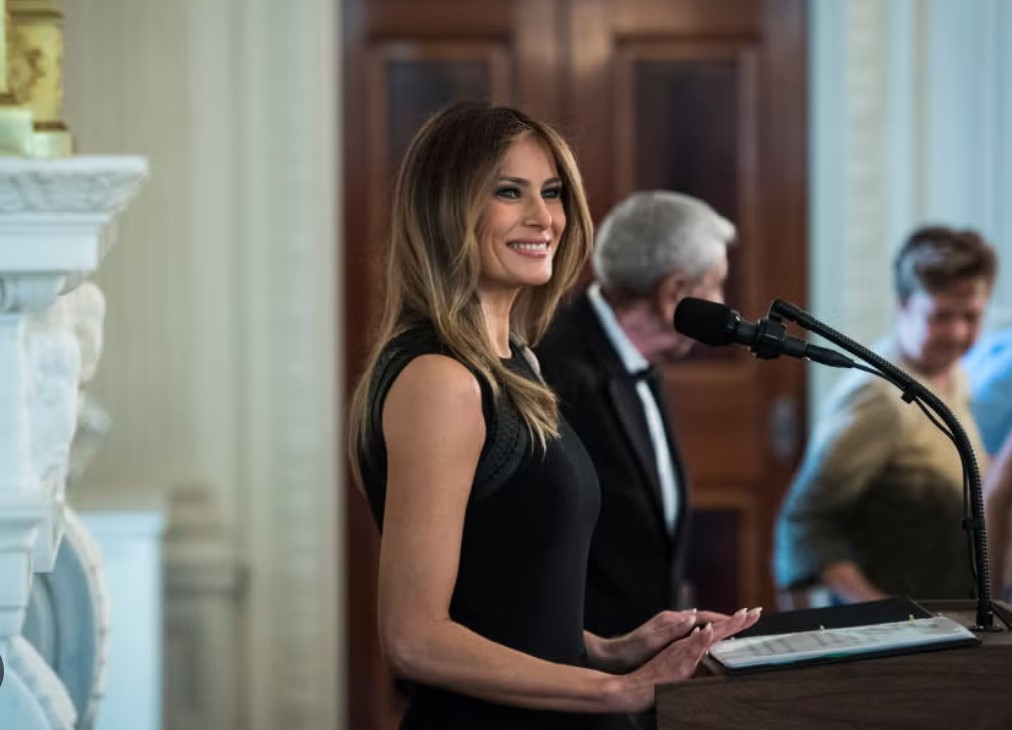 Melania Trump wearing a black dress stands at a podium in a government building